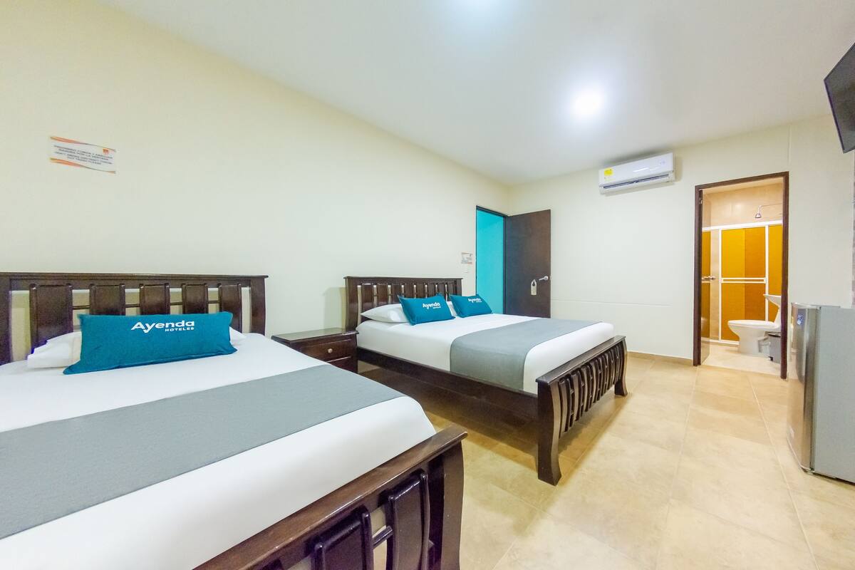 Une chambre d’hôtel avec deux lits, chacun doté d’une tête de lit en bois et d’un oreiller bleu. Une petite table se trouve entre les lits, et une porte donne accès à la salle de bain.