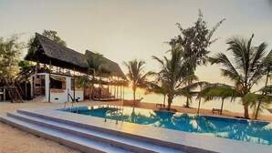 Zona de piscina con escalones que conducen a una estructura sombreada, rodeada de palmeras y un atardecer al fondo.