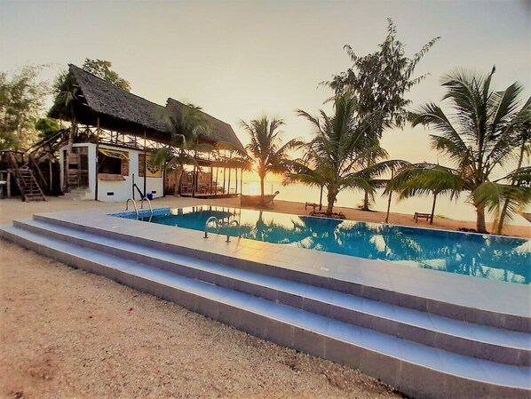 Zona de piscina con escalones que conducen a una estructura sombreada, rodeada de palmeras y un atardecer al fondo.