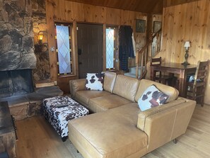 A cozy cabin interior with a stone fireplace, a leather sofa with cowhide cushions, a wooden staircase, and a dining table with chairs.