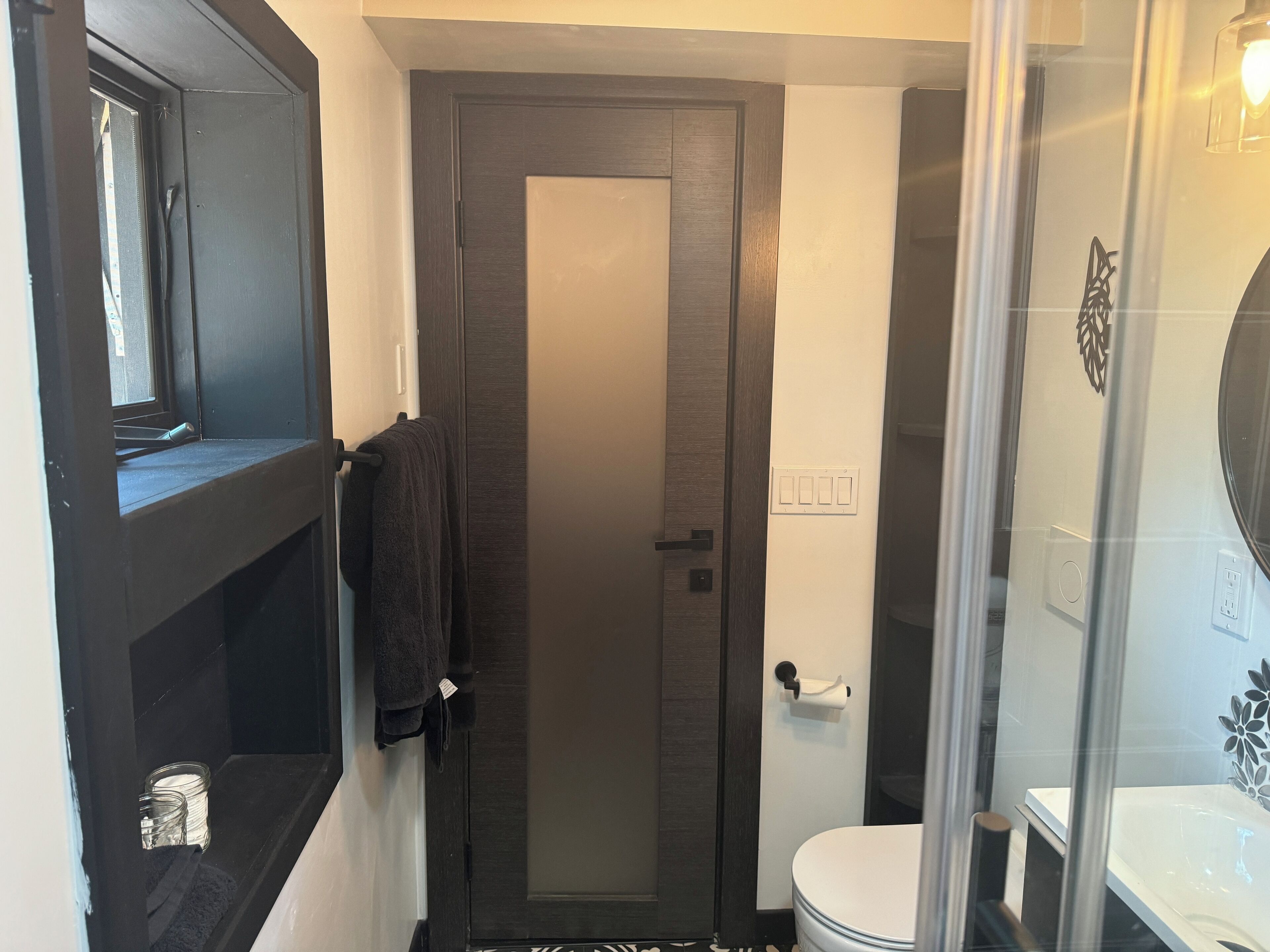 Une salle de bain avec une porte en bois foncé, des toilettes blanches, une cabine de douche en verre et un porte-serviettes sur lequel est accrochée une serviette.