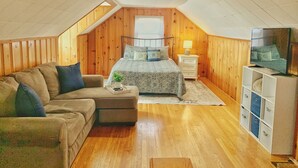 A cozy attic bedroom with a wooden sloped ceiling, a bed with a patterned duvet, a sofa with cushions, a bedside table with a lamp, and a TV on a stand. - Cozy, Convenient, & Comfortable Retreat w/ Firepit, Hammock, Ping Pong, & Coffee (Roanoke)