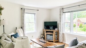 A cozy living room with a sofa, a coffee table, and a TV on a wooden stand. - Cozy, Convenient, & Comfortable Retreat w/ Firepit, Hammock, Ping Pong, & Coffee (Roanoke)