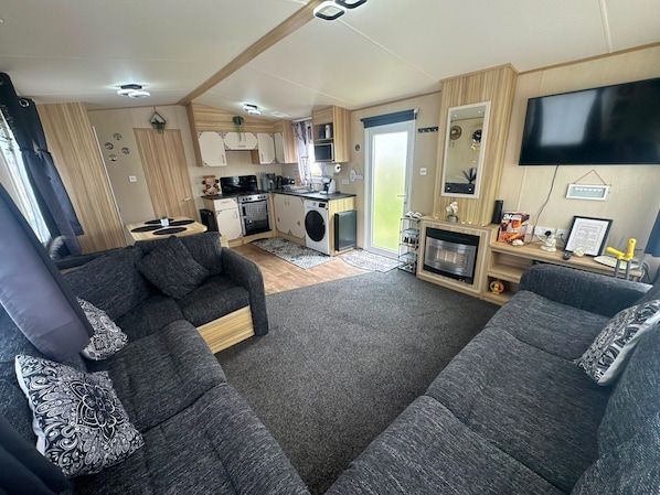 A compact RV interior with a kitchenette, two sofas, a flat-screen TV, and a fireplace.
