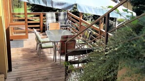 A wooden deck with a table and chairs under a large umbrella, surrounded by a wooden railing and greenery.
