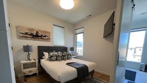 Un dormitorio con cama, mesitas de noche, lámpara, un cuadro y ventana.