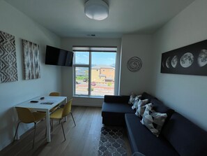 Un soggiorno moderno con un divano, un tavolo da pranzo con sedie, un televisore a schermo piatto montato a parete e una grande finestra con vista sull'esterno.
