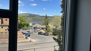 Vista desde una ventana que da a una calle con coches aparcados, cielo azul despejado y un lago al fondo.