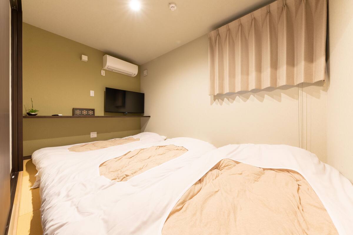 A bedroom with two beds, a flat-screen TV, a small shelf with decorative items, and a window with curtains.
