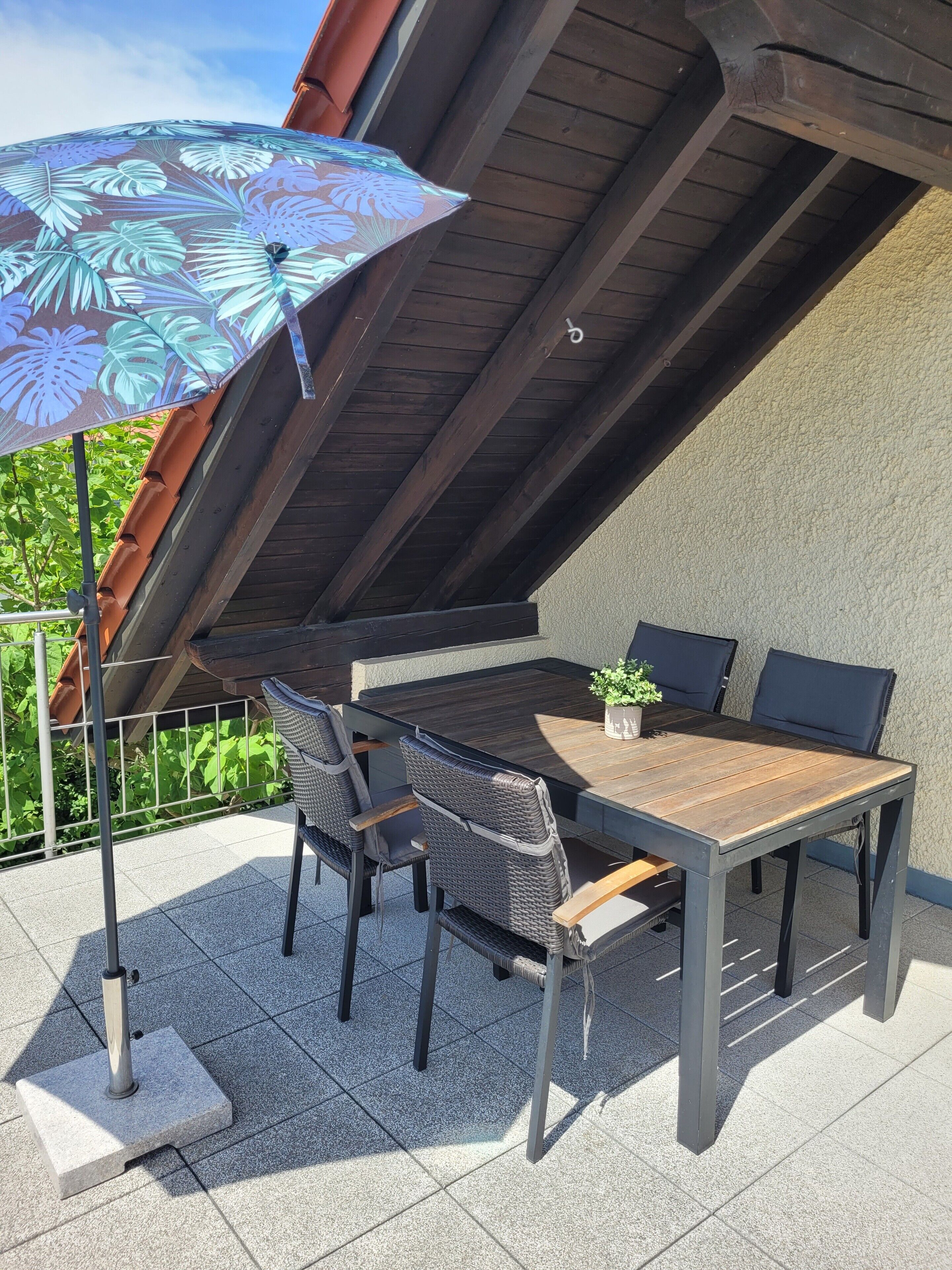 Een terras buiten met een tafel, stoelen, een potplant en een decoratieve parasol.