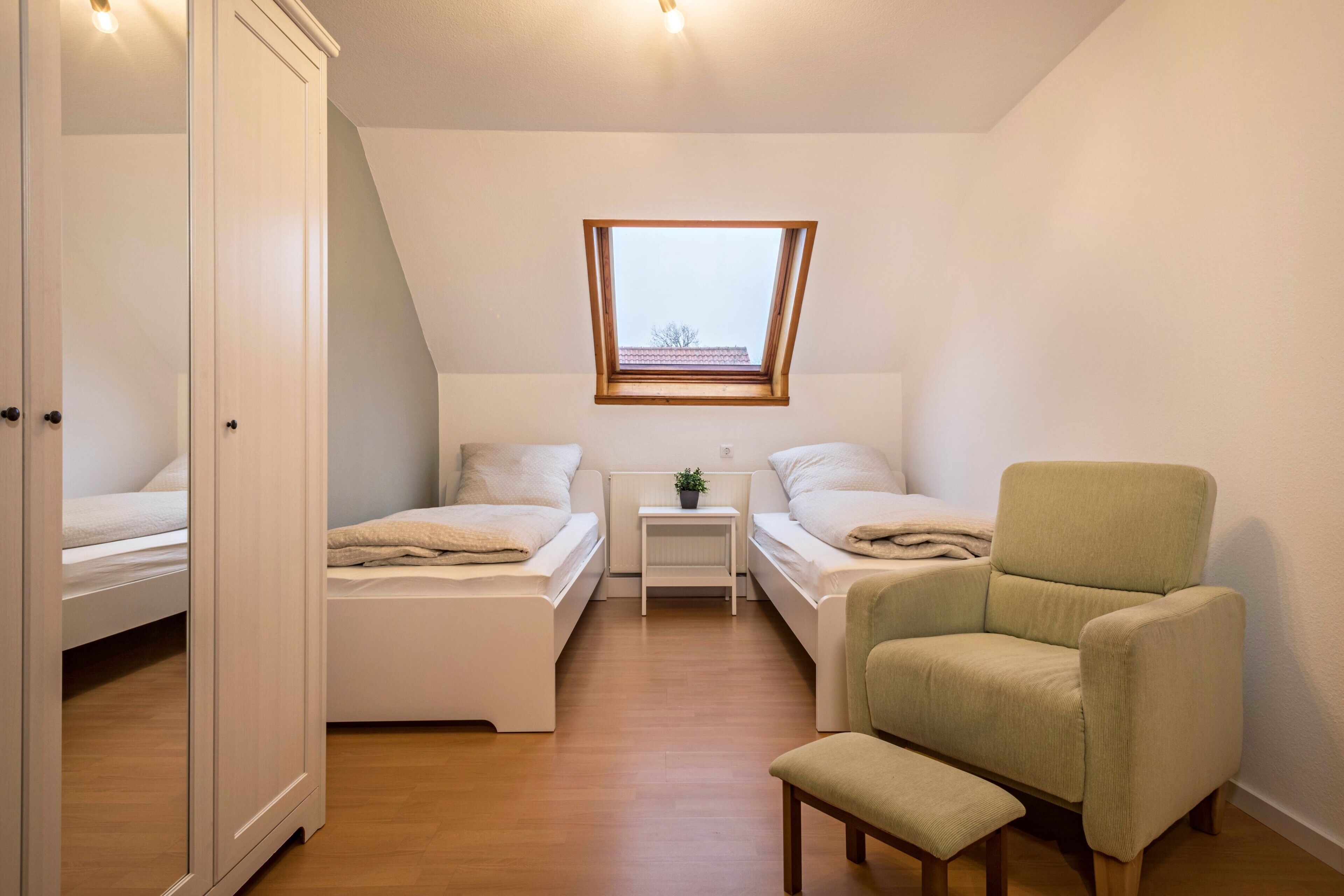A small, well-lit room with two beds, a chair, a small table, and a skylight.