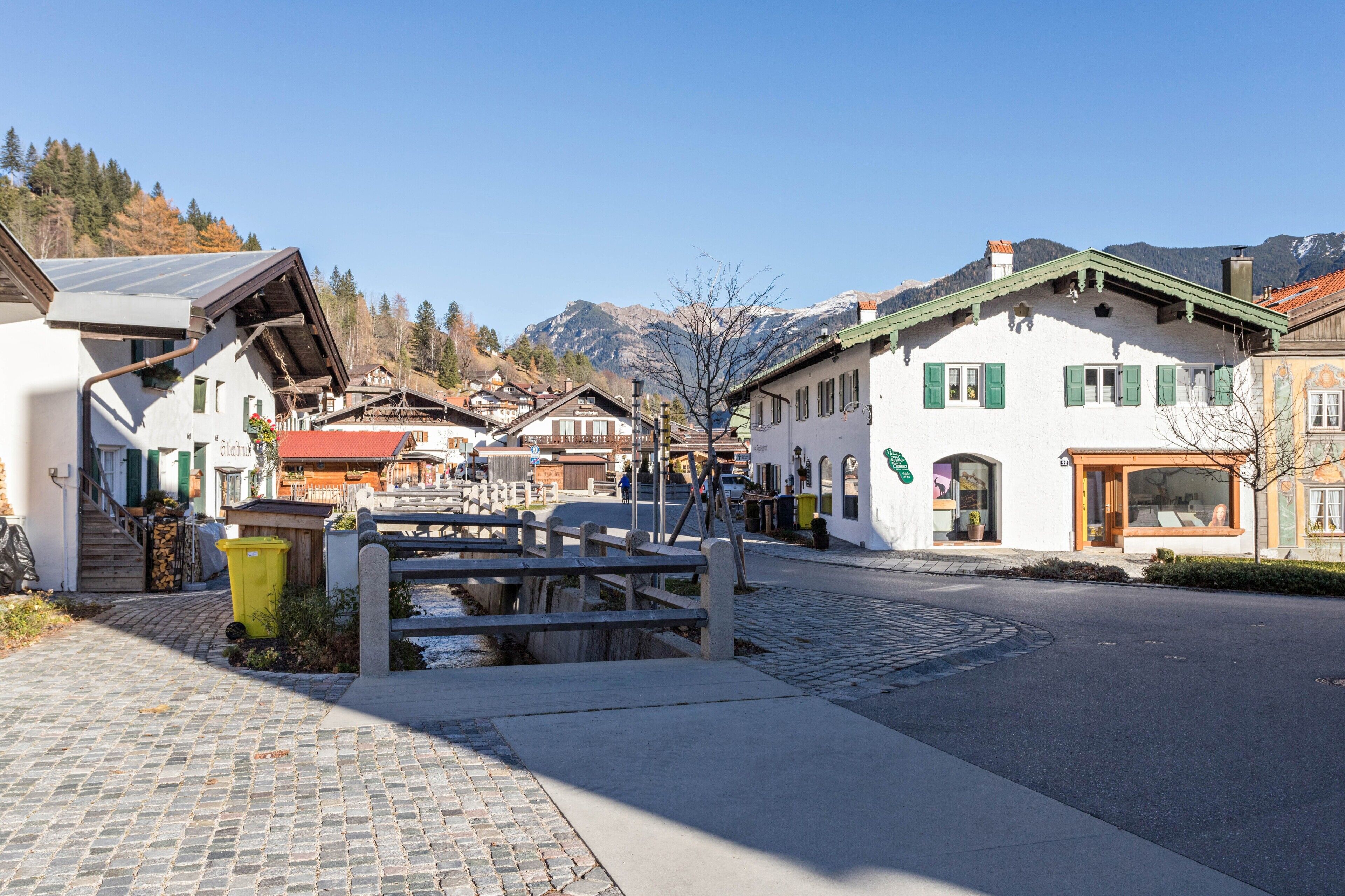 A cobblestone street in a village with traditional buildings, a wooden bridge, and mountains in the background.
