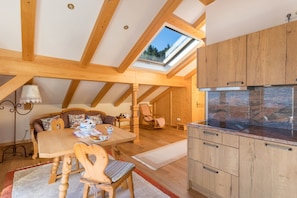 Une pièce mansardée chaleureuse, avec une table à manger en bois, des chaises, un canapé, un fauteuil à bascule, une lucarne et un coin cuisine doté de plans de travail en granit.
