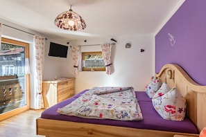 Une chambre à coucher avec un lit, une tête de lit en bois, une fenêtre avec des rideaux, un bureau avec une chaise et un téléviseur fixé au mur.