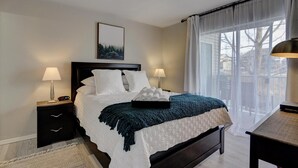 Un dormitorio ordenado con una cama grande, dos mesitas de noche con lámparas, un cuadro en la pared y vistas a través de una ventana con cortinas de encaje.