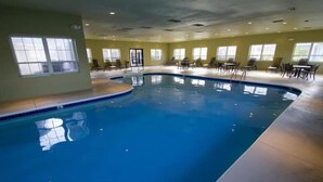 Una piscina interior con agua cristalina, rodeada de sillas de descanso y mesas, y grandes ventanales que permiten la entrada de luz natural.