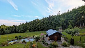A rustic cabin with a stone chimney, surrounded by a well-maintained garden with a pond, pathways, and a dense forest in the background.