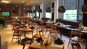 Um restaurante moderno com mesas e cadeiras de madeira, luminárias pendentes pretas e grandes janelas que oferecem vista para o exterior.