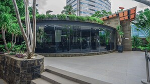 Um edifício moderno com fachada de vidro curva, rodeado por plantas tropicais e palmeiras.
