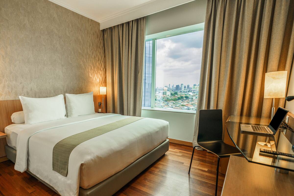 Habitación de hotel con una cama grande, un escritorio con una computadora portátil y vistas a un paisaje urbano a través de un ventanal amplio.