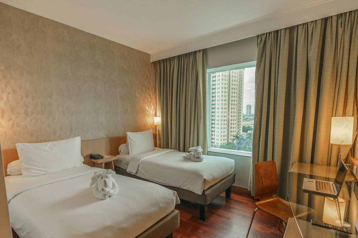 Habitación de hotel con dos camas, una mesita de vidrio con una computadora portátil, una silla y vista a un paisaje urbano a través de grandes ventanales.