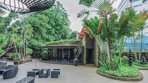 Uma área de estar externa com móveis modernos, cercada por vegetação exuberante e um edifício com telhado de azulejos.