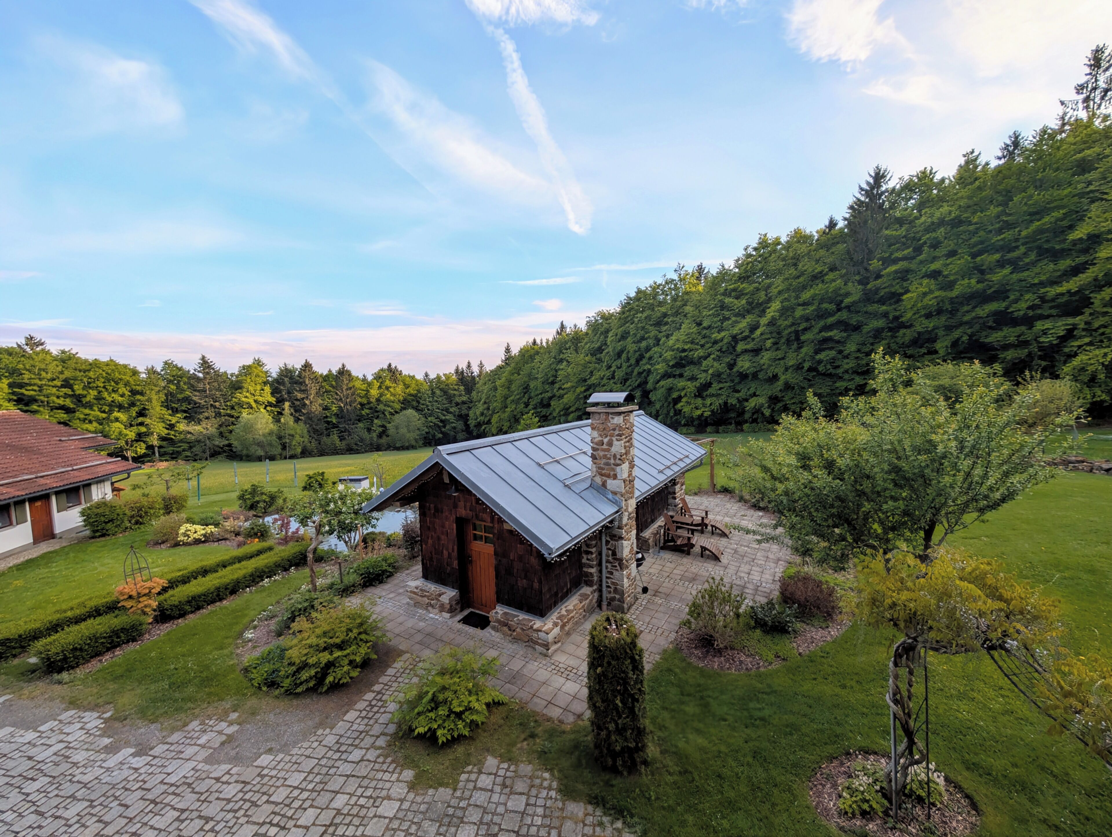 Ein rustikales Blockhaus mit Steinkamin, umgeben von einem gepflegten Garten und Blick auf einen bewaldeten Hügel.