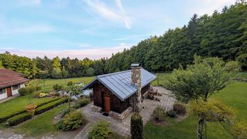 Ein rustikales Blockhaus mit Steinkamin, umgeben von einem gepflegten Garten und Blick auf einen bewaldeten Hügel.