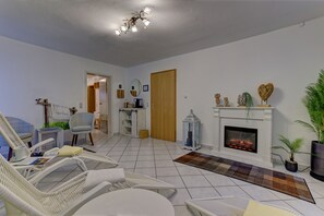 A living room with white walls, a fireplace, a ceiling light fixture, and a door leading to another room.