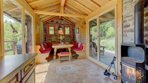 A wooden interior with a stone wall, a fireplace, a table with benches, and a view through large windows.
