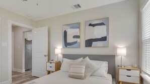 Un dormitorio con una cama, dos mesitas de noche, una mesita auxiliar, una puerta y arte abstracto en la pared.