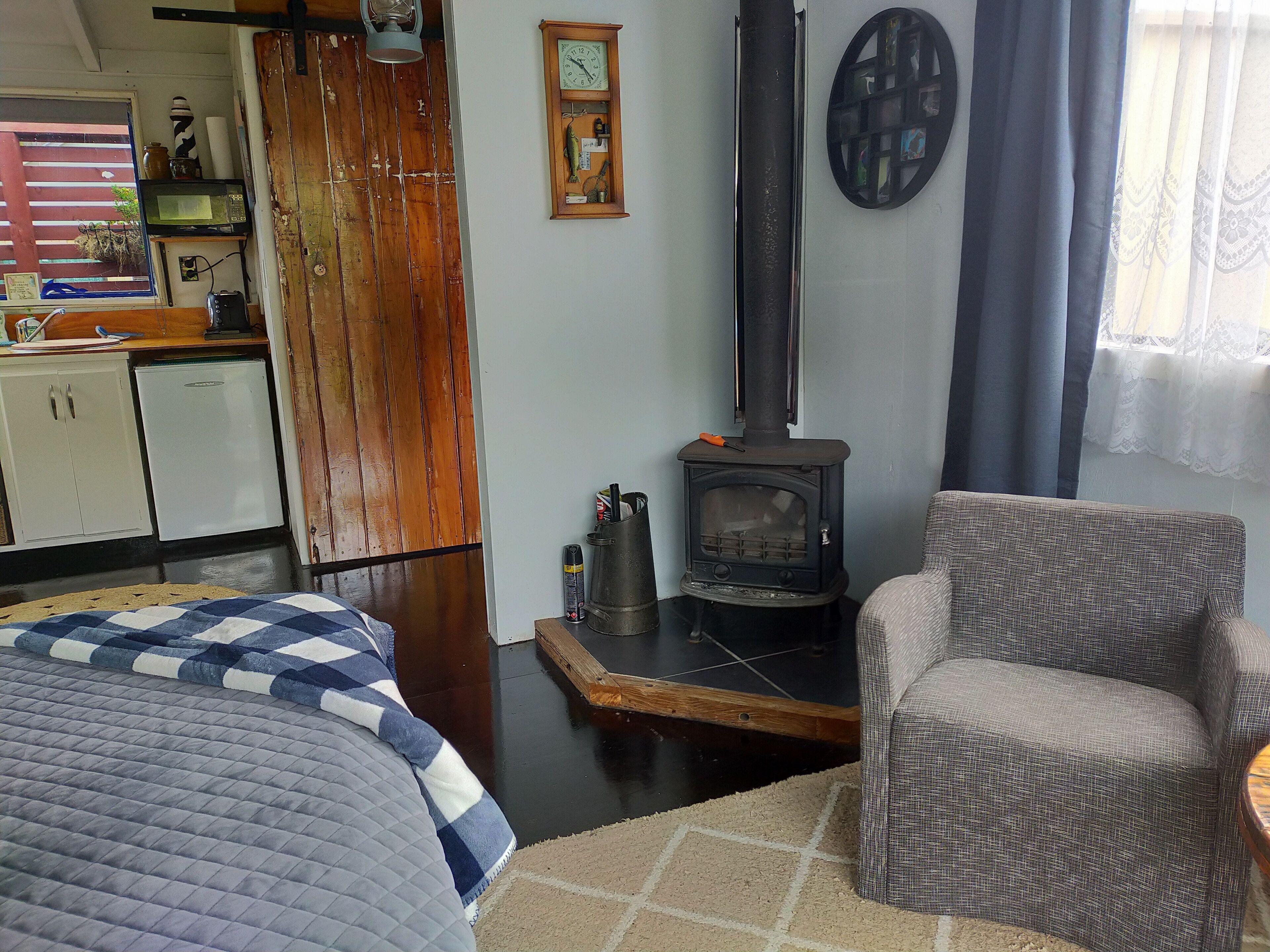 A cozy room with a bed, a chair, a small table, a wooden wardrobe, a microwave, a refrigerator, a clock, a wall-mounted shelf with decorative items, and a wood-burning stove.