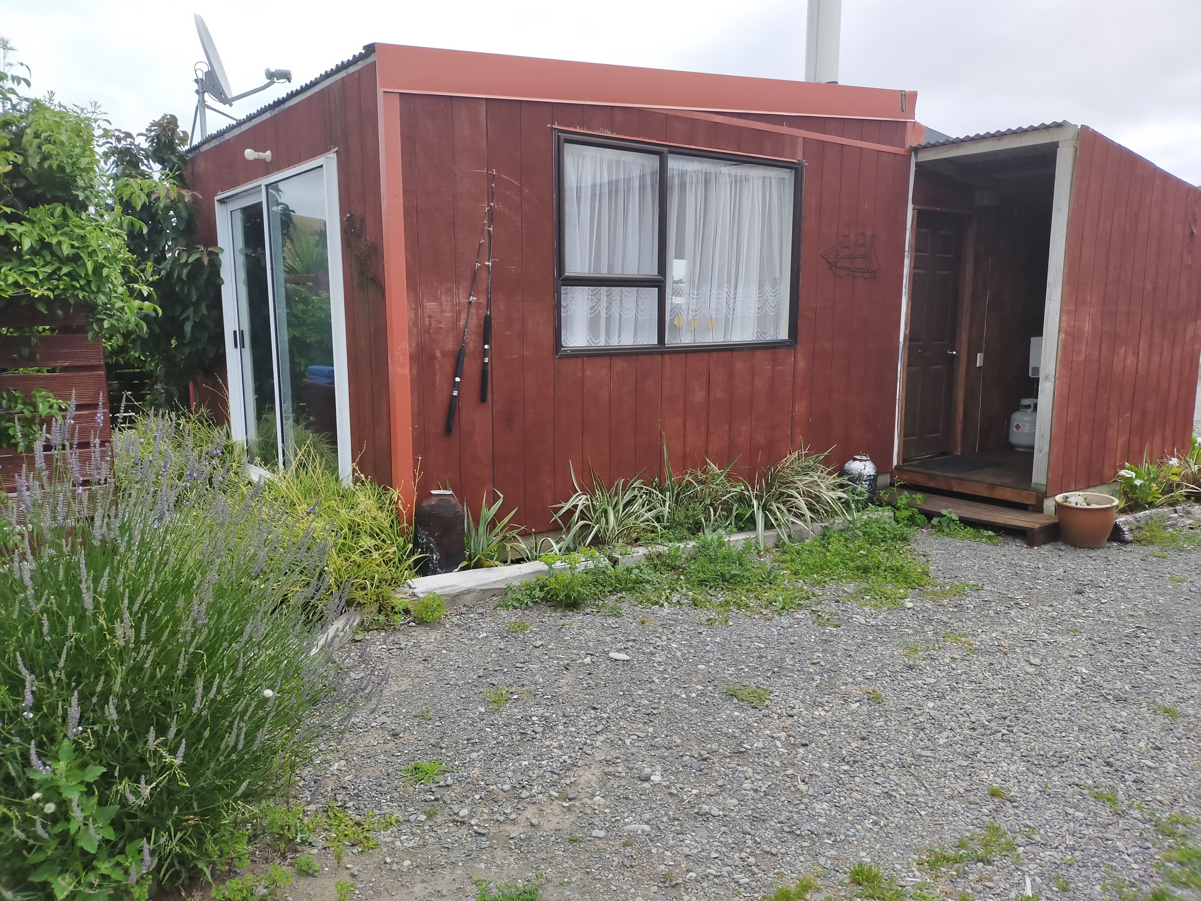 A small, single-story wooden building with a glass door and window, surrounded by gravel and plants.