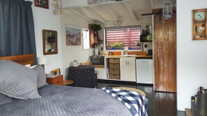 A bedroom with a bed, a small desk, a window with blinds, and a kitchenette area with a microwave, toaster, and sink.