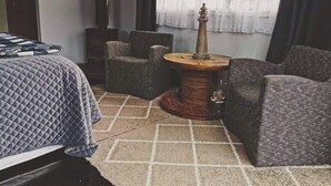 A room with a bed, two armchairs, a small round table with a lamp, and a window with sheer curtains.