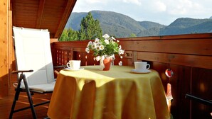 Eine Holzterrasse mit Tisch für zwei Personen, einem weißen Stuhl und Blick auf Berge.