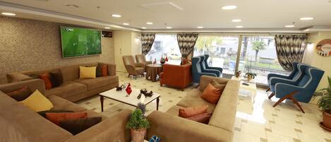 A modern hotel lobby with a large TV screen, comfortable seating, a coffee table with decorative items, and a view of the street through large windows.