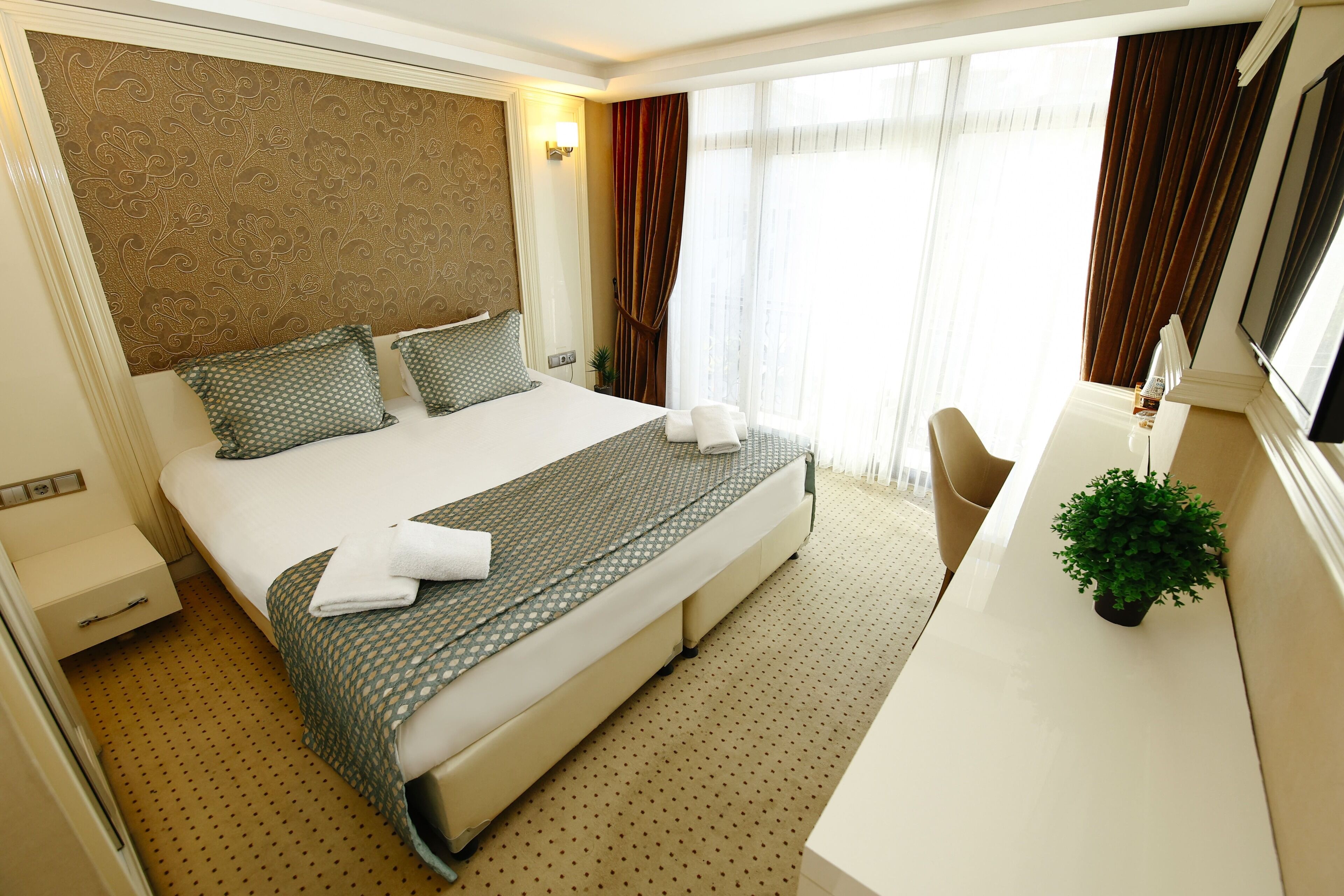 A hotel room with a large bed, a desk, a chair, a potted plant, and a window with curtains.