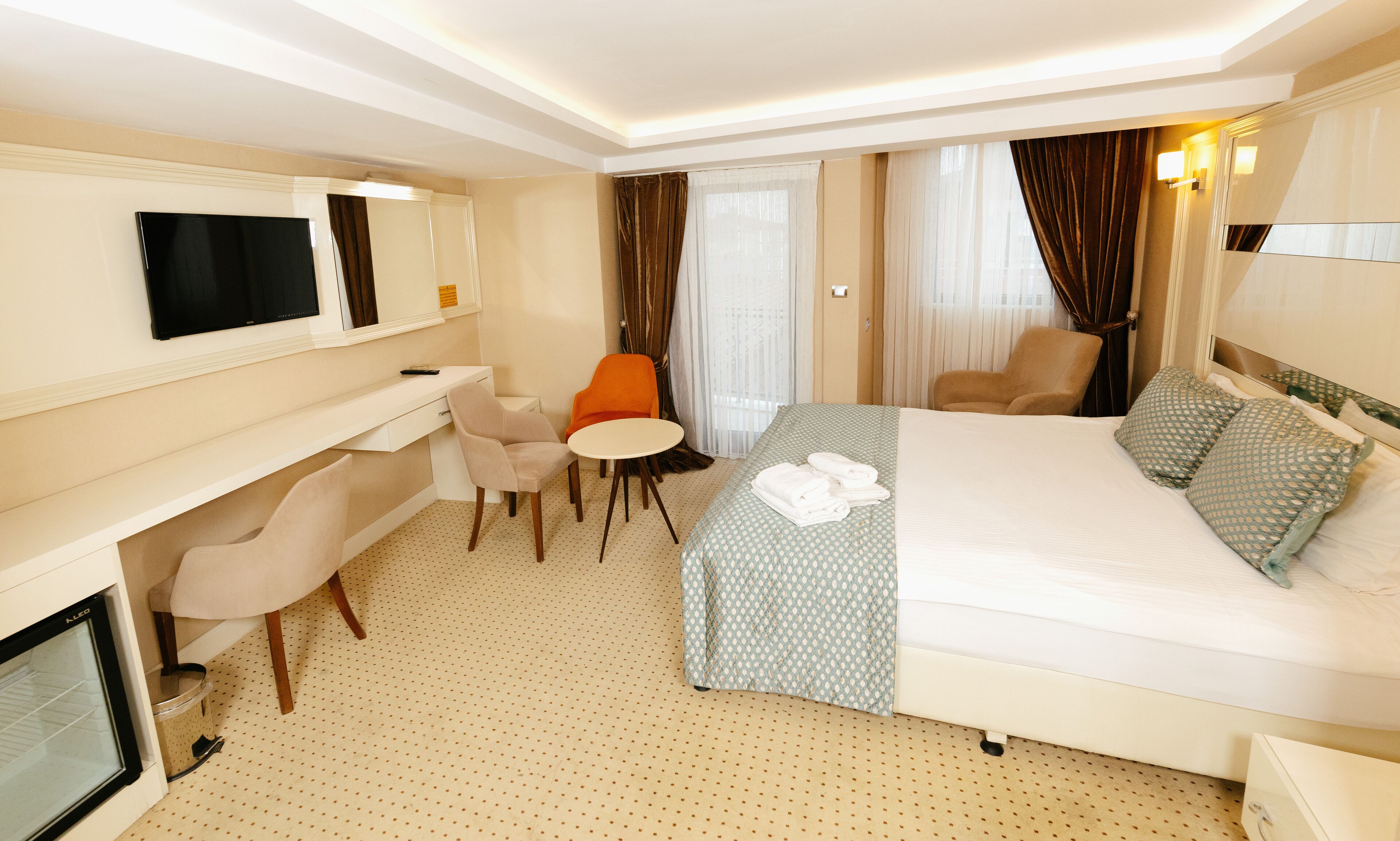 A hotel room with a bed, a desk, an orange chair, a TV, and a window with curtains.