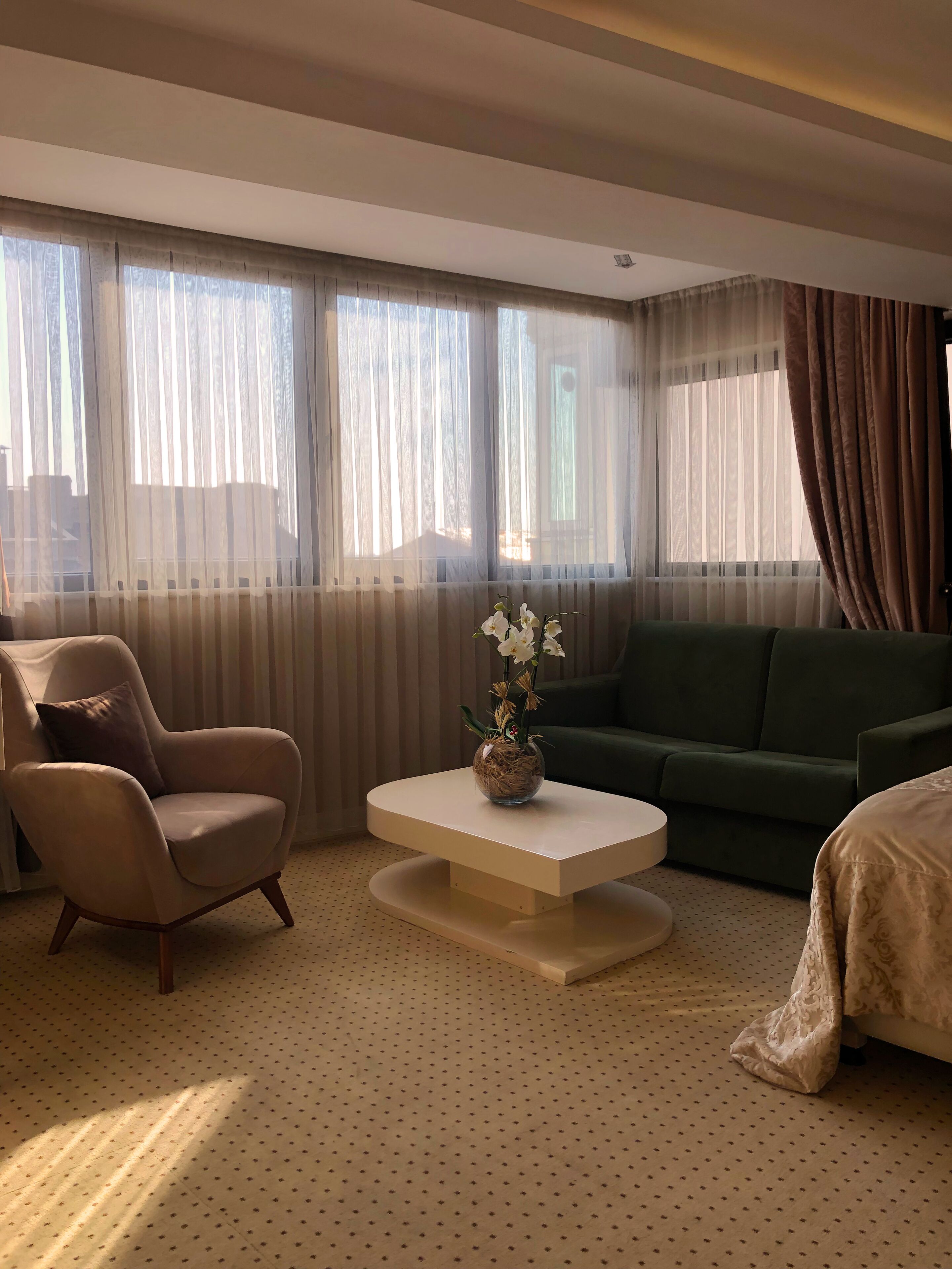 A hotel room with a beige armchair, a green sofa, a white coffee table with a flower vase, and large windows with sheer curtains.