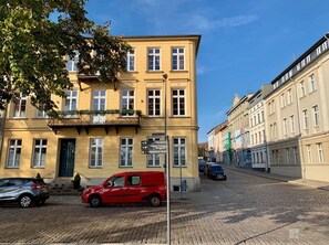 Ein mehrstöckiges Gebäude mit gelber Fassade, einem gut erkennbaren Eingang und einem Schild mit Wegweisungen zu umliegenden Orten.