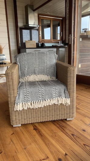 A wicker armchair with a patterned throw blanket, positioned on a wooden floor in a room with a kitchen in the background.