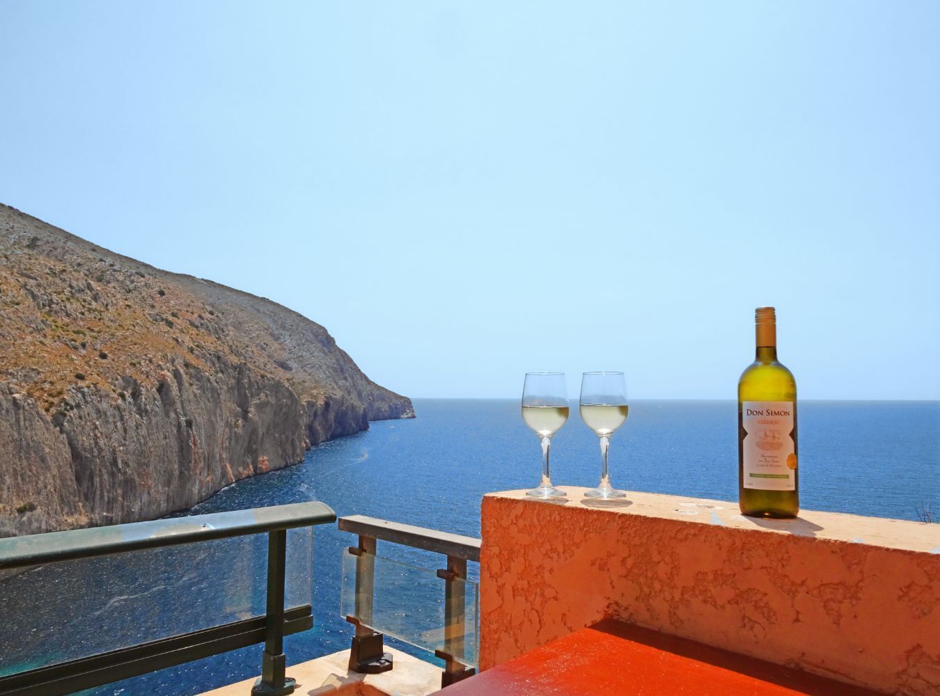 A coastal view with a bottle of Don Simon wine and two glasses on a balcony.