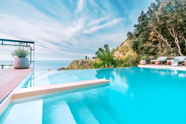 A swimming pool with a view of the sea, surrounded by greenery and a deck with lounge chairs.