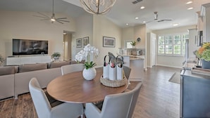 A modern living room with a wooden dining table, white chairs, a sofa, and a TV.