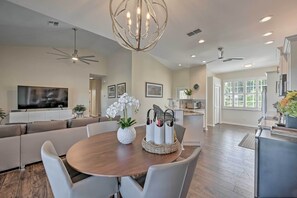 A modern living room with a wooden dining table, white chairs, a sofa, and a TV.