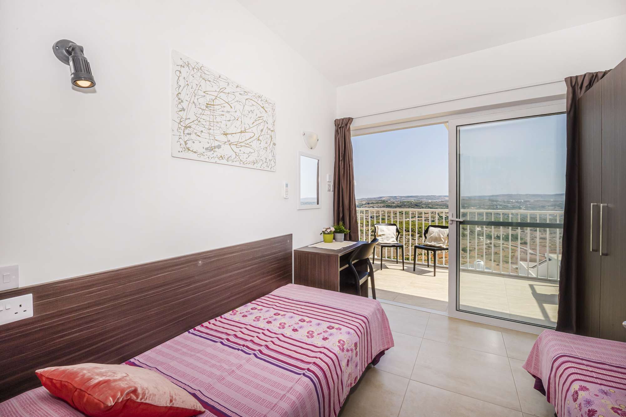 Quarto de hotel com duas camas, uma mesa, uma cadeira, varanda com vista e um quadro na parede.