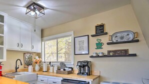 Eine Küche mit weißen Schränken, einer Kaffeemaschine und einem Kühlschrank. An der Wand befinden sich Dekorationen und ein Fenster mit Jalousien.