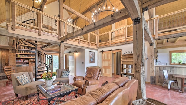 A spacious living area with a high ceiling, wooden beams, a spiral staircase, leather sofas, a coffee table, and a large window. - Bloomington Cabin: 4 Mi to Indiana University (Bloomington)