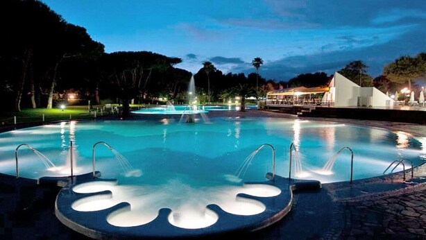 A circular pool with a central fountain, surrounded by a paved area with steps leading to a covered seating area.